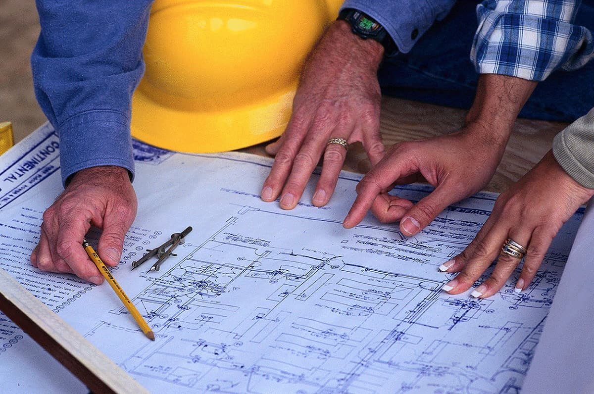 Three people's hands gathered around a large blue architectural blueprint - two male hands and one female hand pointing at different details on the blueprint