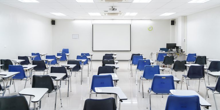 Empty classroom with blue and black student desks arranged neatly, white projection screen in front
