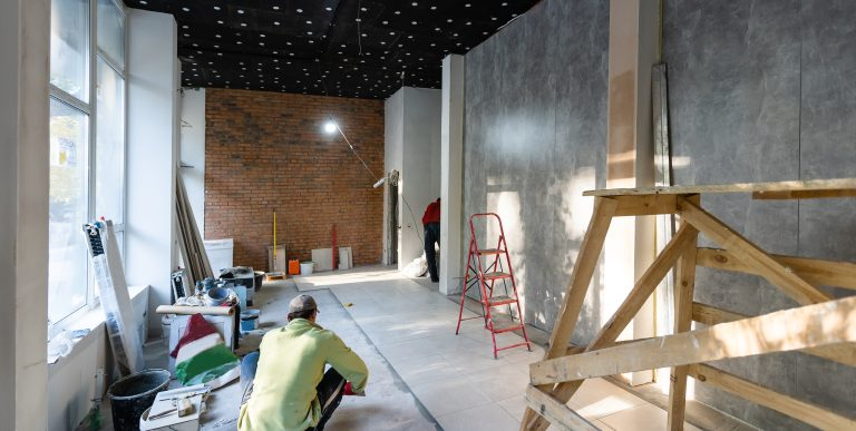 Construction worker in green shirt and hat working on floor installation, red brick wall and gray panel wall visible