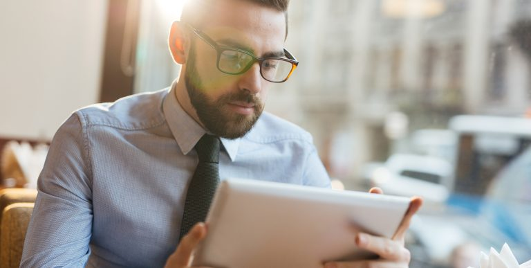 Bearded man in blue shirt and tie looking down at white tablet computer with bright sunlight