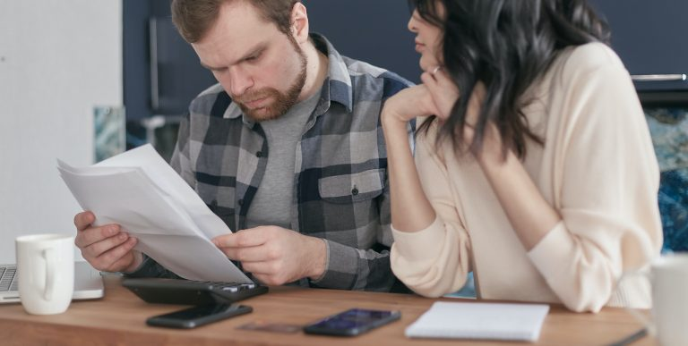 Bearded man in plaid shirt reviewing documents with woman, calculator and smartphone on desk